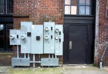 A view from the sidewalk of a cluster of electricity meters by a metal door in the side of a brick building.