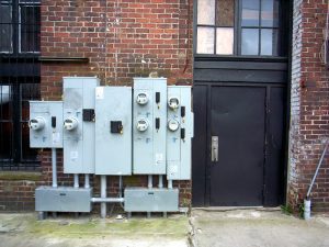 A view from the sidewalk of a cluster of electricity meters by a metal door in the side of a brick building.