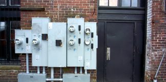 A view from the sidewalk of a cluster of electricity meters by a metal door in the side of a brick building.