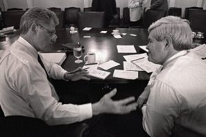A black-and-white image of President Clinton and Newt Gingrich seated at a table. Clinton, at left, is gesturing with his right hand. On the gleaming wooden table are several pieces of paper and goblets of water.