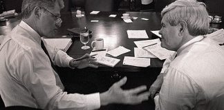 A black-and-white image of President Clinton and Newt Gingrich seated at a table. Clinton, at left, is gesturing with his right hand. On the gleaming wooden table are several pieces of paper and goblets of water.