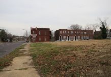 View of an empty city lot. At the far end is a row of brick apartment houses.