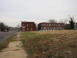 View of an empty city lot. At the far end is a row of brick apartment houses.