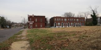 View of an empty city lot. At the far end is a row of brick apartment houses.