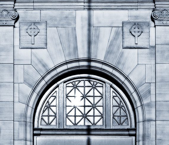 Black-and-white close-up of church architecture, showing arched window over a door and bas-relief crosses higher up.