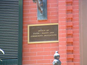 A bronze plaque on an exterior red-brick wall, with a dark green shutter at left. The plaque reads "Office of Faith-Based and Community Initiatives. "