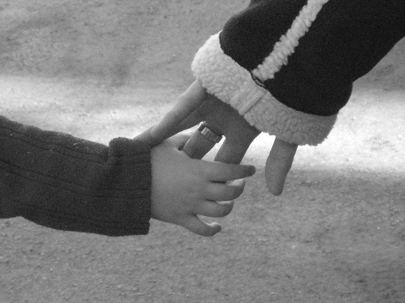 Close-up view from behind of two hands, a child's hand holding two fingers of a woman's hand. Both are wearing coats.