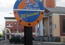 A round blue and orange sign on an urban street corner with a train on it reads "Welcome to the Ironbound"