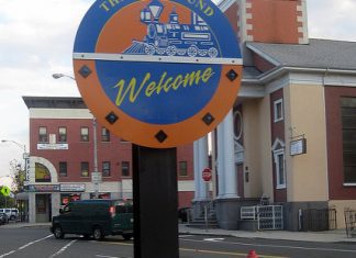 A round blue and orange sign on an urban street corner with a train on it reads "Welcome to the Ironbound"