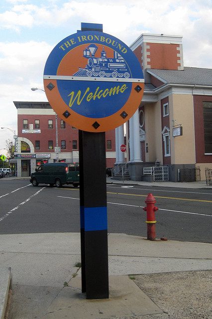 A round blue and orange sign on an urban street corner with a train on it reads "Welcome to the Ironbound"