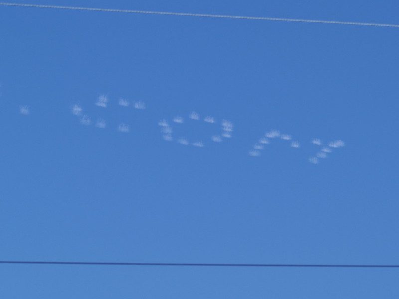 Against a blue sky, skywriting planes have spelled out ".com" as if by dot matrix printer, for an article on community-labor relations in the post dot-com boom
