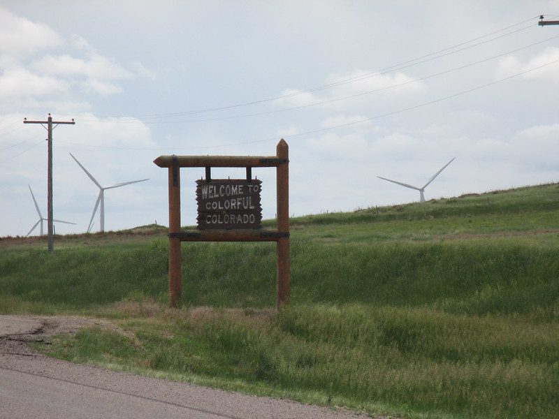 A rugged wooden sign announces "Welcome to Colorful Colorado" against a hillside with windmills, for an article on smart growth in Colorado