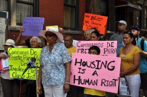 A group of about a dozen people, of varying ages and colors, in summer clothes, stand on the sidewalk holding homemade posters in support of a rent strike. A young person holds poster that reads "Housing is a human right." 