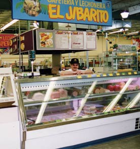 A glass-fronted display case at El Mercado, a project of Bickerdike Redevelopment. The proprietor, a young man in a ball cap, stands behind the counter with his forearms resting on the counter. Above him is suspended a blue sign with yellow lettering: "El Jibarito"