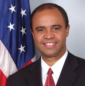 A formatl headshot of Adolfo Carrion, with the American flag just behind his right shoulder. He's wearing a suit and red tie, and is smiling.