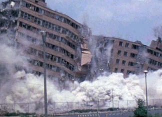 A high-rise public housing complex being demolished. The upper stories are beginning to collapse as smoke and rubble rises up from the ground