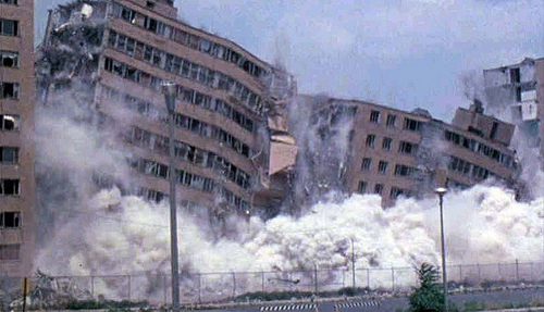 A high-rise public housing complex being demolished. The upper stories are beginning to collapse as smoke and rubble rises up from the ground