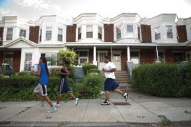 Photo shows three young African Americans jogging on a wide sidewalk in front of brown rowhouses with white trim, and hedges along the walk. Illustrating article about health philanthropy
