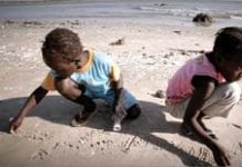 Two children, one wearing a blue shirt and other a pink shirt, play in the sand on a beach.