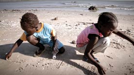 Two children, one wearing a blue shirt and other a pink shirt, play in the sand on a beach.
