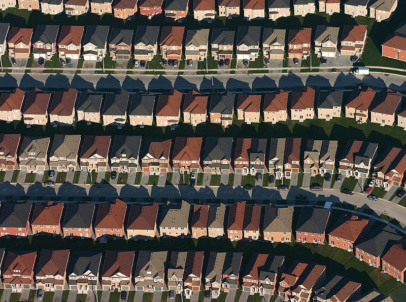 An aerial view of a suburban development shows six rows of close-set, very similar-looking houses, for an article examining homeownership as the path to wealth for Americans of color. Where you can see the house fronts, there are one or two cars in the driveways. The yards are small and there aren't any trees. 