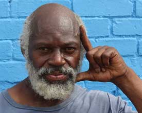 Close up of an African-American Vietnam veteran with white beard standing in front of a bright blue walk, and holding his hand up to the left side of his face in an "L" shape.