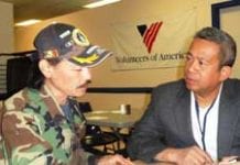 A man in camouflage jacket an hat talks at a table with a man in a suit, with a Volunteers of America banner on the wall behind them.