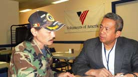 A man in camouflage jacket an hat talks at a table with a man in a suit, with a Volunteers of America banner on the wall behind them.