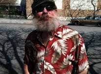 Navy veteran Mark Hudgins, a white man in a black knit hat with a white beard and a red shirt with a white feather print looks at the camera, standing outside.