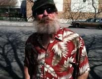 Navy veteran Mark Hudgins, a white man in a black knit hat with a white beard and a red shirt with a white feather print looks at the camera, standing outside.