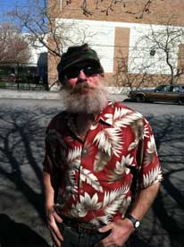 Navy veteran Mark Hudgins, a white man in a black knit hat with a white beard and a red shirt with a white feather print looks at the camera, standing outside.