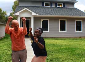 A man and woman express joy with raised arms and big smiles on the walk leading to a gray house. The man is light skinned and bald-headed and is wearing an orange shirt and tan pants. The woman is dark skinned and wearing a black top, light tan skirt, and has a headband in her hair. She looks poised to jump. The house has a white portico over the front door and three vertical windows with white frames and dark glass. The front yard is green grass.