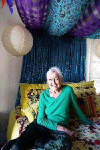 A smiling woman with gray hair and a green top sits on a bed covered with colorful throw pillows, with blue and purple fabric hanging on the back wall and from the ceiling.