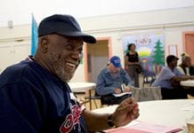 A smiling African-American man in a baseball cap sits at a table in a social lounge type room with other people in the background.
