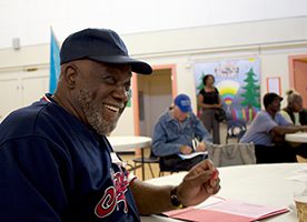 A smiling African-American man in a baseball cap sits at a table in a social lounge type room with other people in the background.