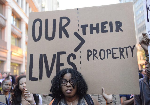 A woman holds a cardboard sign that reads "Our Lives > Their Property"