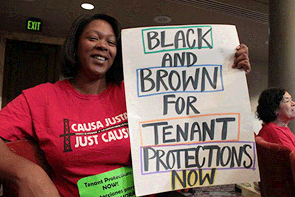A woman in a red Cause Justa/Just Cause T-shirt holds a hand-lettered sign reading "Black and Brown for Tenant Protections Now"