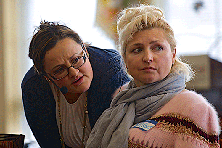 Two women, one on the right stares off camera as the other bends to speak to her.