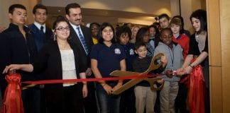 A group of people stand behind a red ribbon before the opening of an art exhibit at a charter school. To the right, youngsters prepare to cut the ribbon with large scissors.