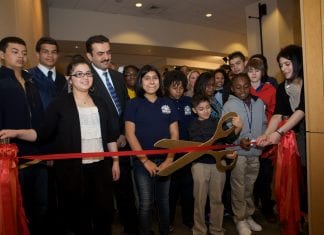 A group of people stand behind a red ribbon before the opening of an art exhibit at a charter school. To the right, youngsters prepare to cut the ribbon with large scissors.