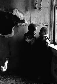A black-and-white photo of two boys by a window, in front of a greatly deteriorated wall, with exposed pipes and possibly lead paint dust or chips.