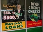 A sign for payday loans is seen on a window, with a illustration of a man in a suit holder cash in his arms. The sign also reads "Fast friendly service. $50 to $500."