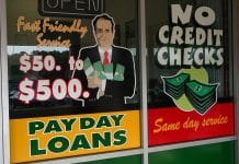 A sign for payday loans is seen on a window, with a illustration of a man in a suit holder cash in his arms. The sign also reads "Fast friendly service. $50 to $500."