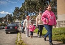 A woman in a pink, "I love San Francisco" sweater walks ahead of two women in light beige sweaters and a little girl who is wearing a pink coat and drinking a bottle of water. They are walking down a street and passing a black car that is parked on the left.