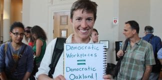 A woman holds a sign outside of the Oakland City Council chambers that reads "Democratic Workplaces equal Democratic Oakland!"
