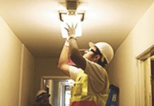 A trainee wearing a white hardhat fixes a light fixture in an apartment.