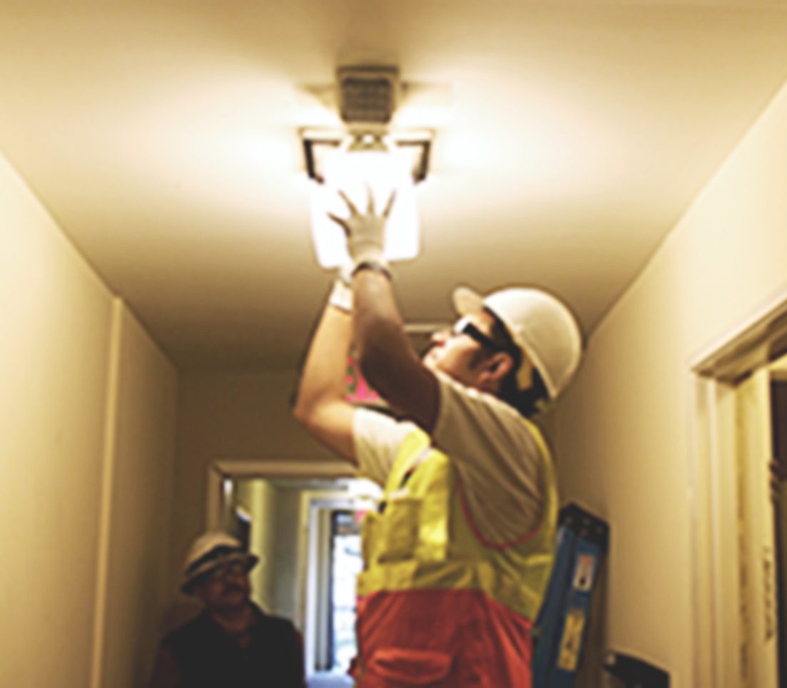 A trainee wearing a white hardhat fixes a light fixture in an apartment.