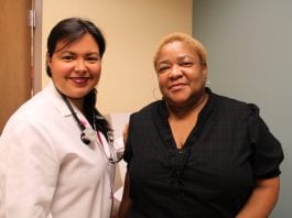 A female doctor t the Daughters of Charity Health Center in New Orleans wears a white lab coat with a stethoscope and stands next to an African American woman, who is wearing a black shirt.