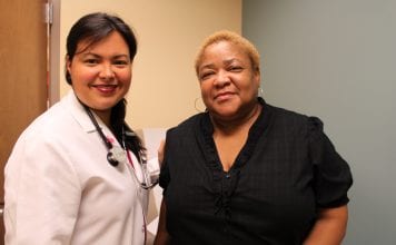 A female doctor t the Daughters of Charity Health Center in New Orleans wears a white lab coat with a stethoscope and stands next to an African American woman, who is wearing a black shirt.
