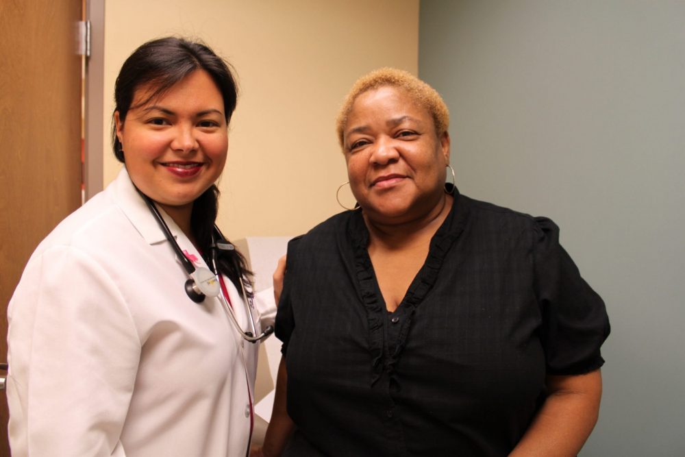 A female doctor t the Daughters of Charity Health Center in New Orleans wears a white lab coat with a stethoscope and stands next to an African American woman, who is wearing a black shirt.